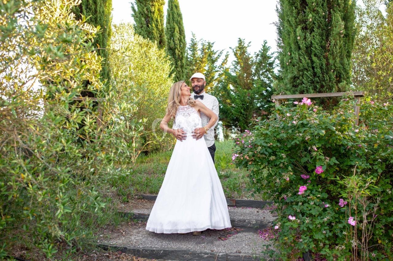 Un couple en tenue de mariage pose dans un jardin verdoyant entouré de fleurs et de cyprès.