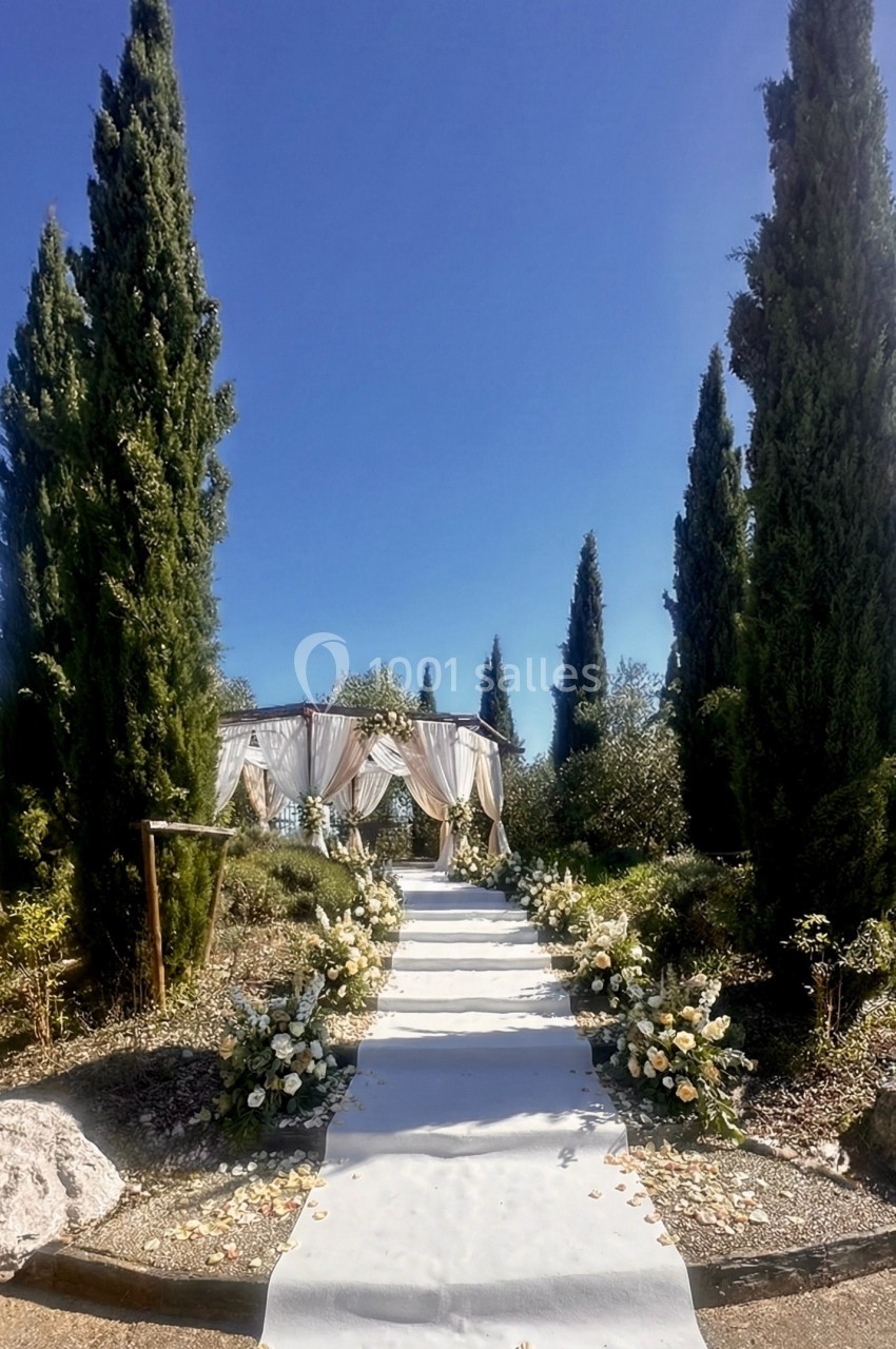 Allée décorée de fleurs menant à une arche de mariage ornée de voilages, entourée de cyprès sous un ciel bleu.