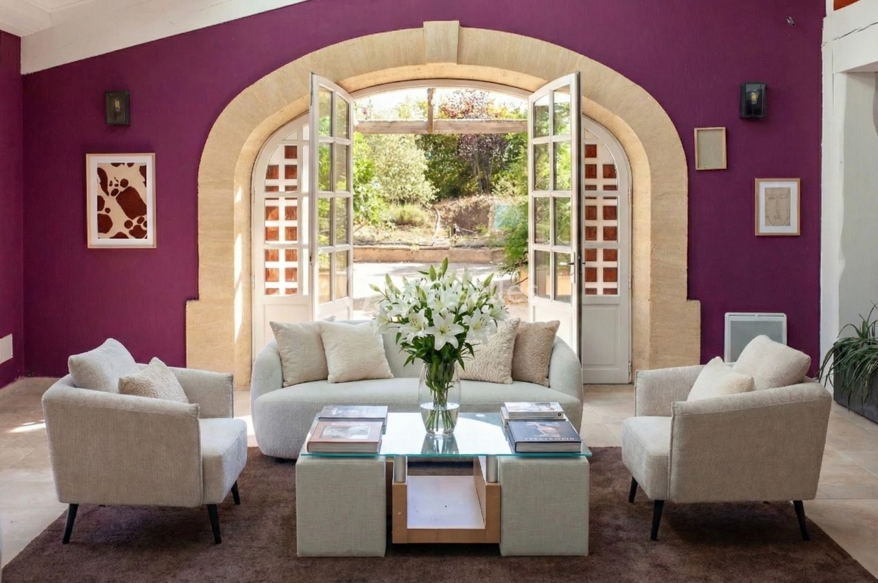 Salon lumineux avec murs violets, canapé et fauteuils beiges, table basse avec fleurs blanches, vue sur jardin par des…