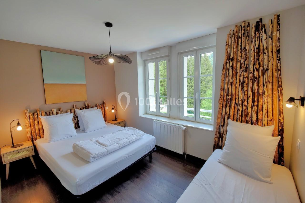 Location salle Crozets (Jura) - Domaine Le Coupet #8 Chambre lumineuse avec deux lits simples et un lit double, parquet sombre, grandes fenêtres et décoration naturelle.