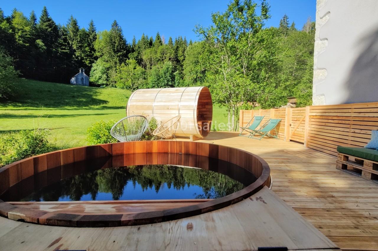 Location salle Crozets (Jura) - Domaine Le Coupet #3 Terrasse en bois avec bain nordique, sauna en tonneau, chaises longues et vue sur une prairie bordée de forêt.