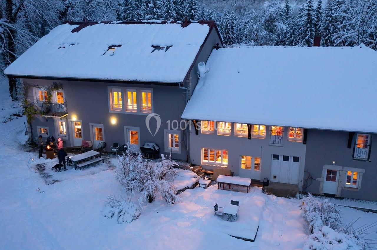 Location salle Crozets (Jura) - Domaine Le Coupet #5 Vue aérienne d'une maison enneigée avec des lumières allumées, entourée d'arbres en hiver.