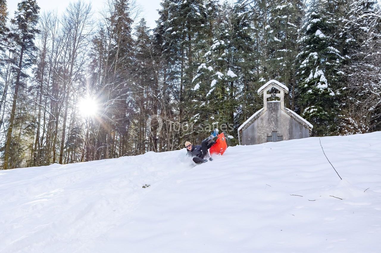 Location salle Crozets (Jura) - Domaine Le Coupet #15 Une personne descend une pente enneigée sur une luge près d'une petite chapelle entourée d'arbres.