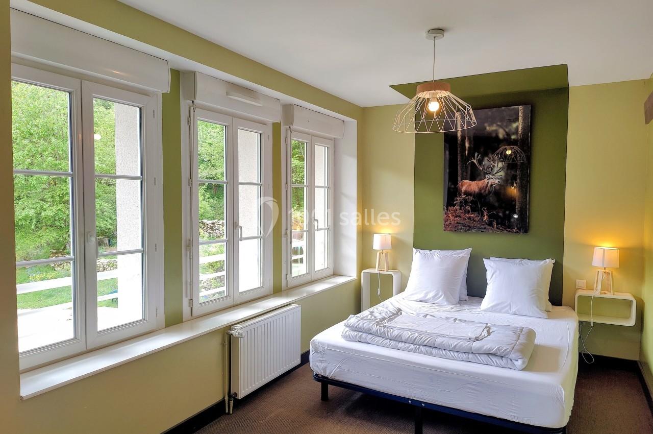 Location salle Crozets (Jura) - Domaine Le Coupet #9 Chambre lumineuse avec lit double, grandes fenêtres donnant sur un paysage verdoyant et décor minimaliste.