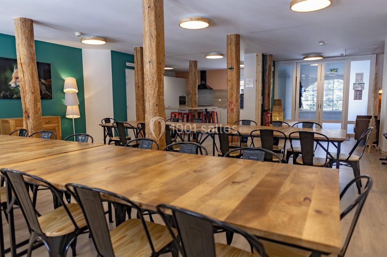 Location salle Crozets (Jura) - Domaine Le Coupet #6 Salle lumineuse avec de grandes tables en bois, chaises métalliques, colonnes en bois et cuisine ouverte en arrière-plan.