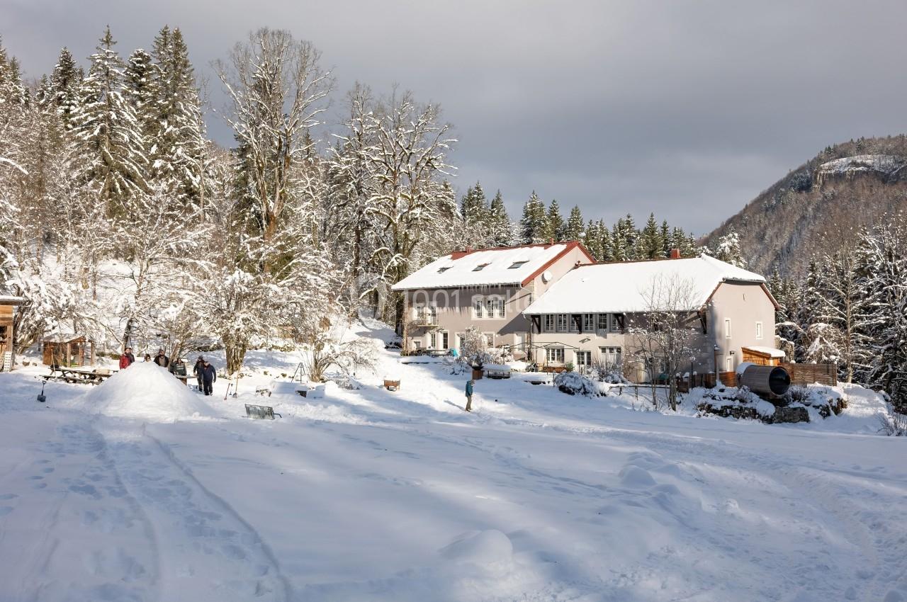 Location salle Crozets (Jura) - Domaine Le Coupet #17 Paysage enneigé avec des bâtiments entourés d'arbres, des personnes profitant de la neige au premier plan.