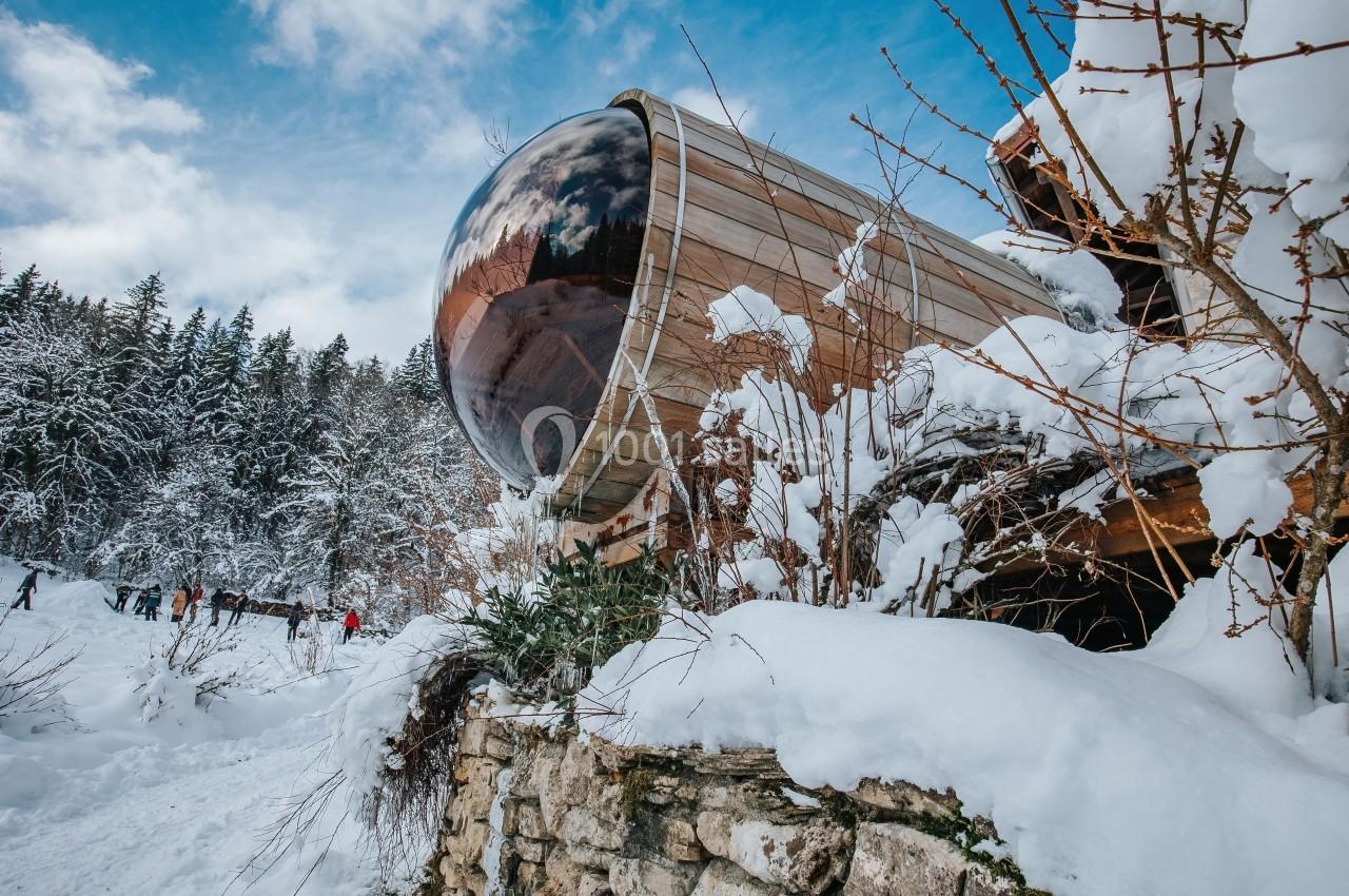 Location salle Crozets (Jura) - Domaine Le Coupet #18 Cabane en bois avec une grande fenêtre arrondie, entourée de neige et de végétation hivernale, près d'une forêt.