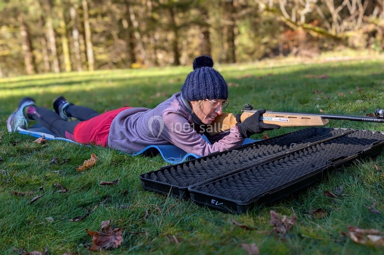 Location salle Crozets (Jura) - Domaine Le Coupet #20 Une personne allongée sur l'herbe vise avec une carabine, un étui ouvert posé à côté, dans un environnement boisé.