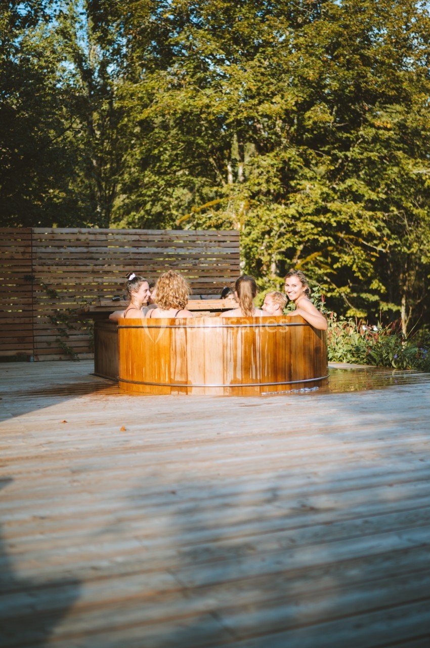 Location salle Crozets (Jura) - Domaine Le Coupet #4 Des personnes se détendent dans un bain nordique en bois, entouré de végétation et d'une terrasse en bois.