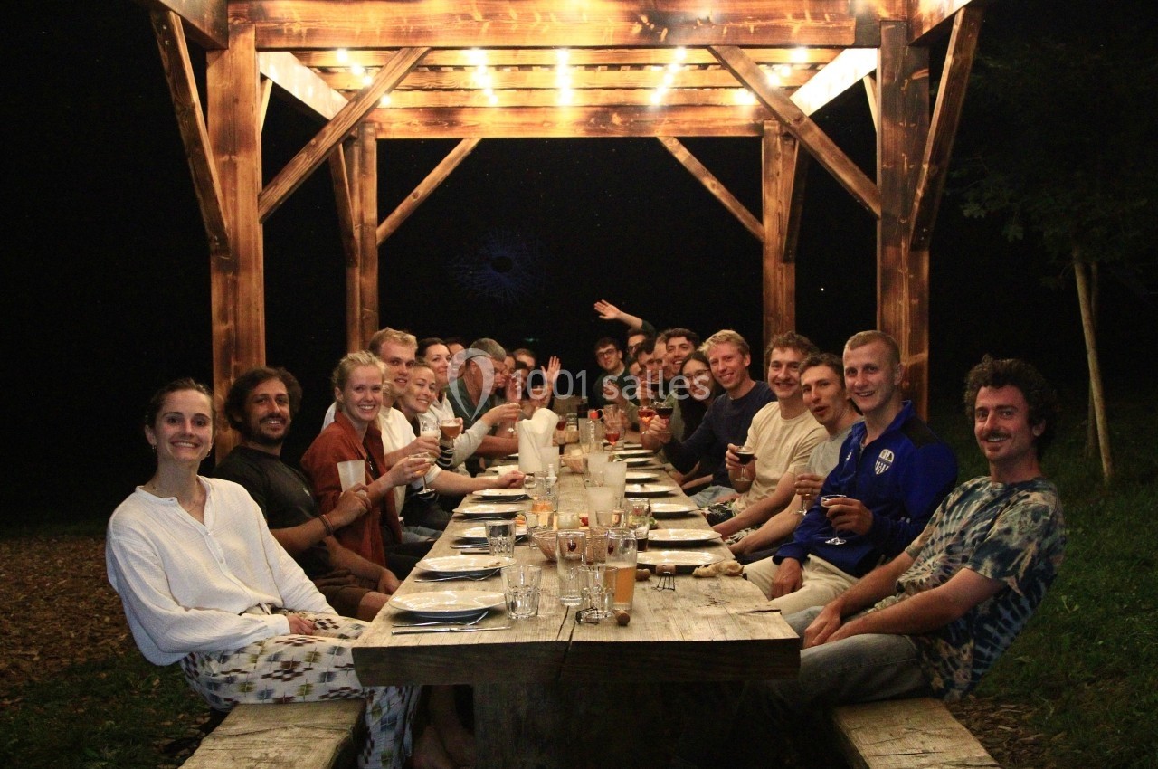 Location salle Crozets (Jura) - Domaine Le Coupet #23 Un groupe de personnes assises autour d'une longue table en bois sous une pergola éclairée, de nuit.