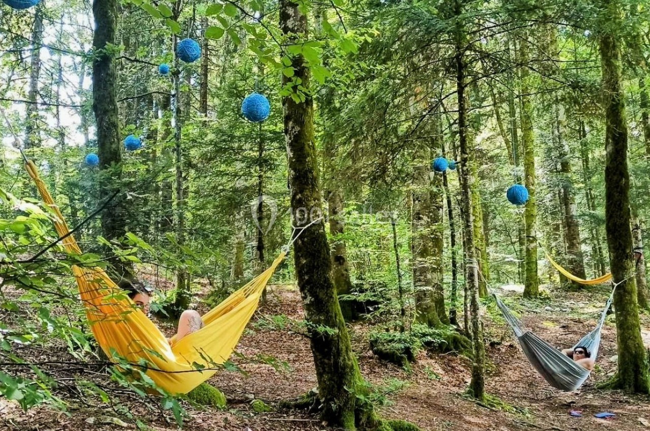 Location salle Crozets (Jura) - Domaine Le Coupet #24 Des personnes se reposent dans des hamacs colorés suspendus entre des arbres dans une forêt décorée de lanternes bleues.