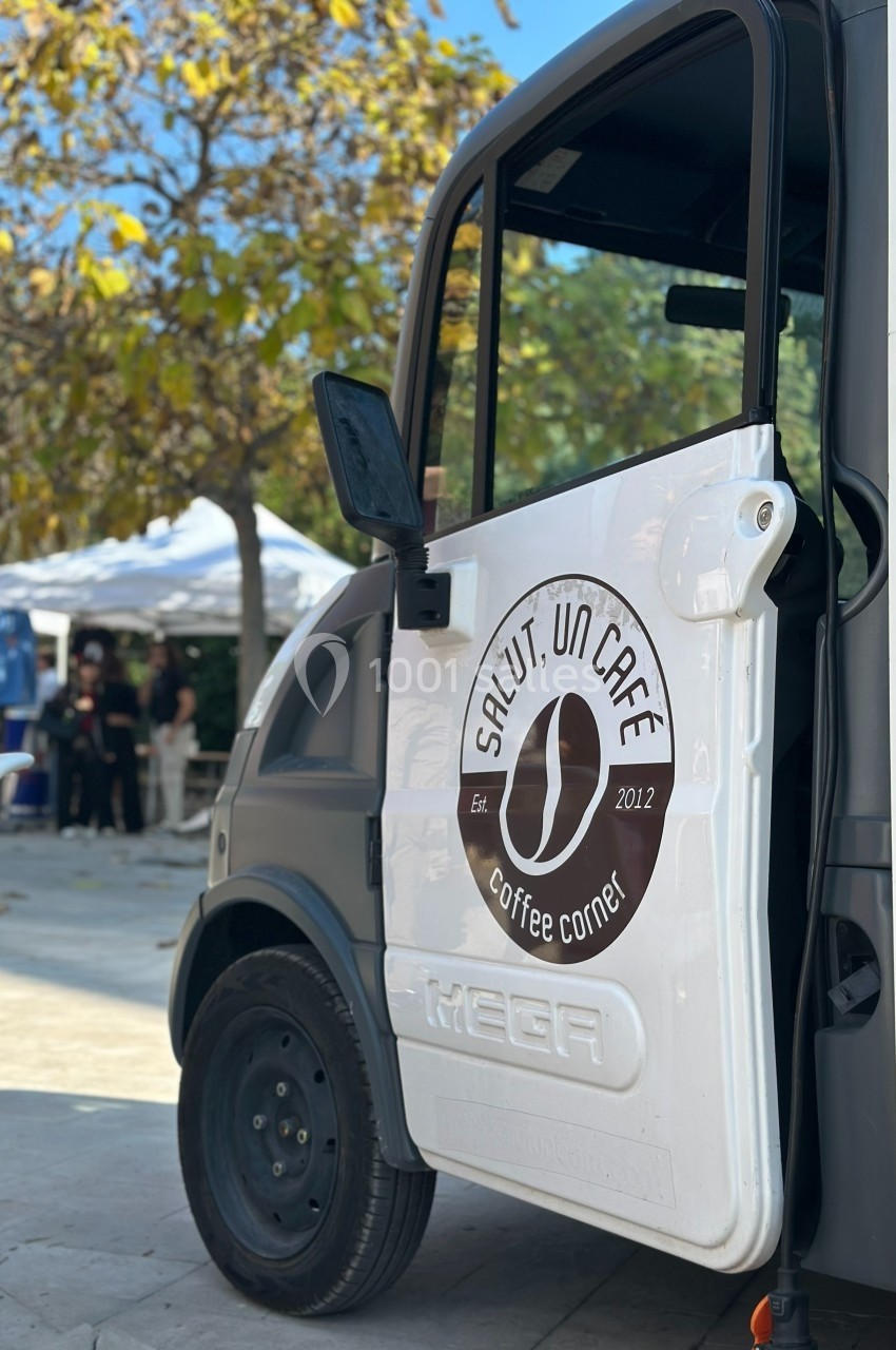 Camionnette de café avec une porte ouverte, stationnée dans un espace extérieur bordé d'arbres et de stands.