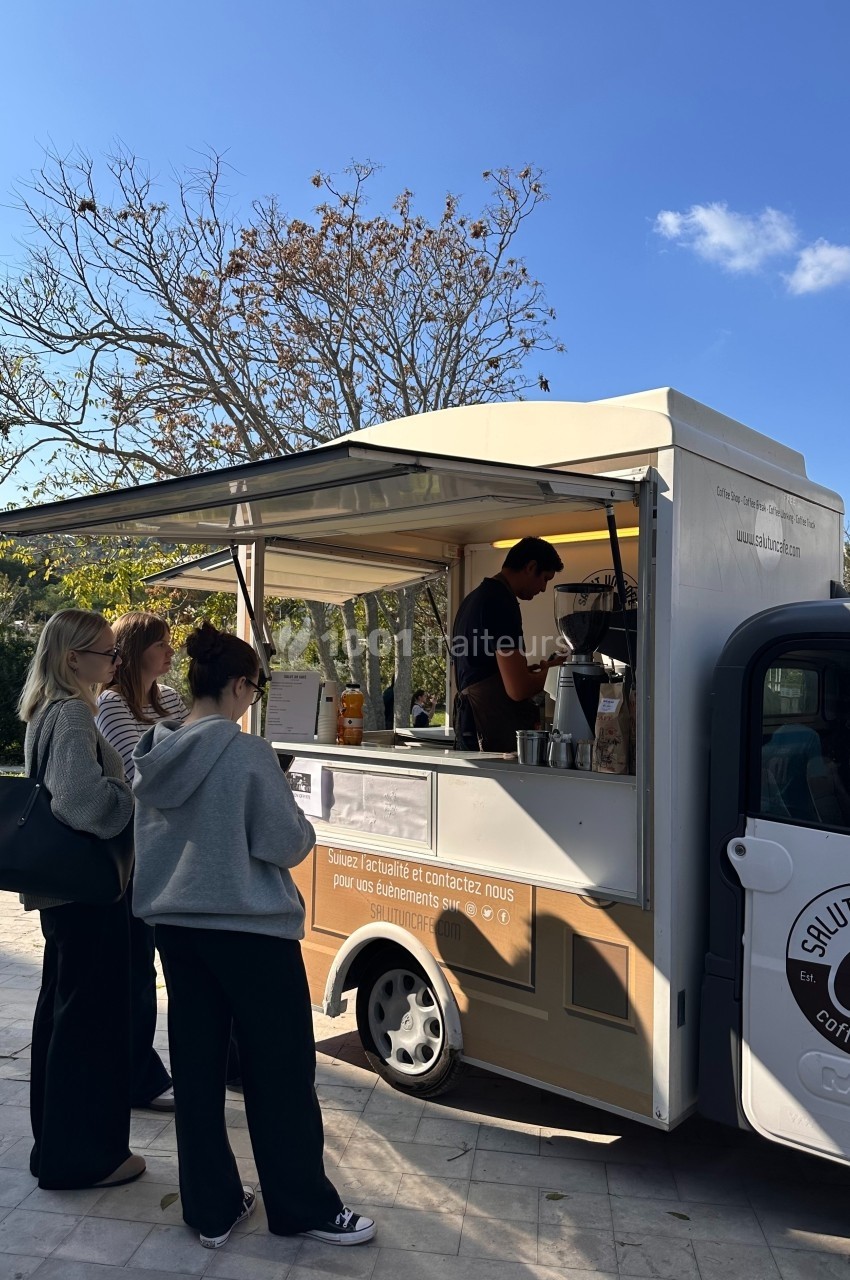 Un food truck sert des boissons à trois clientes en plein air, sous un ciel dégagé avec des arbres en arrière-plan.
