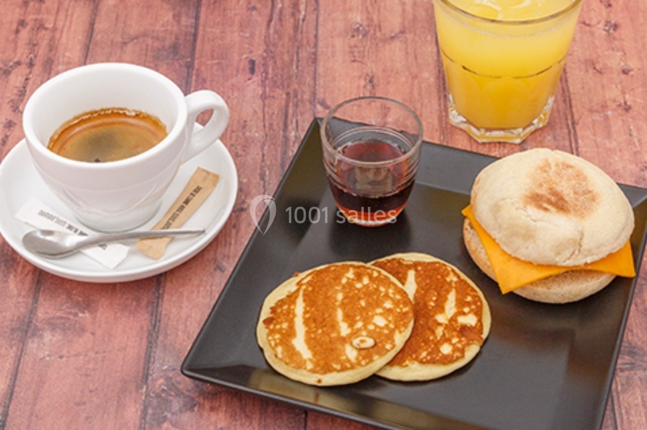Petit-déjeuner composé de pancakes, muffin au fromage, café, jus d'orange et sirop sur une table en bois.