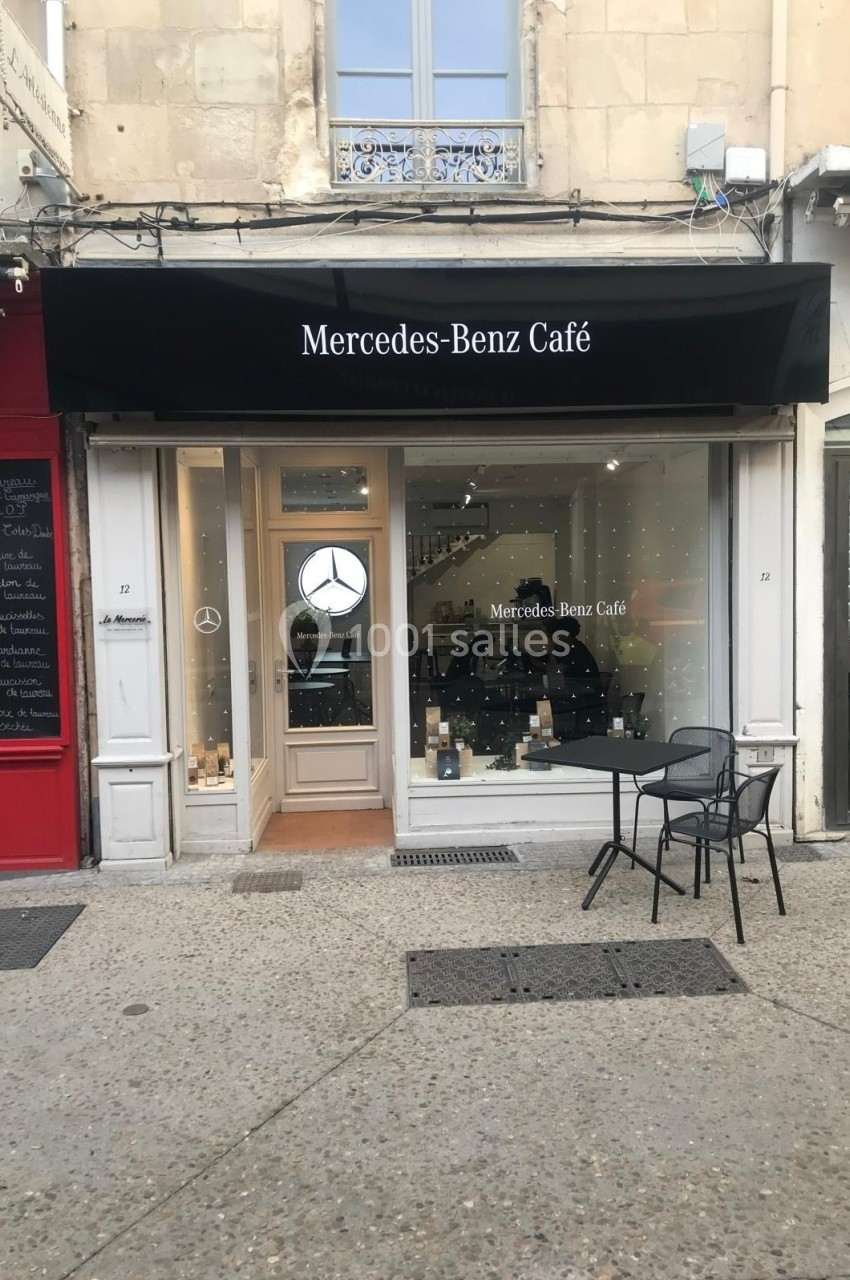 Façade d'un café avec enseigne ’Mercedes-Benz Café’, terrasse avec une table et deux chaises sur un trottoir.