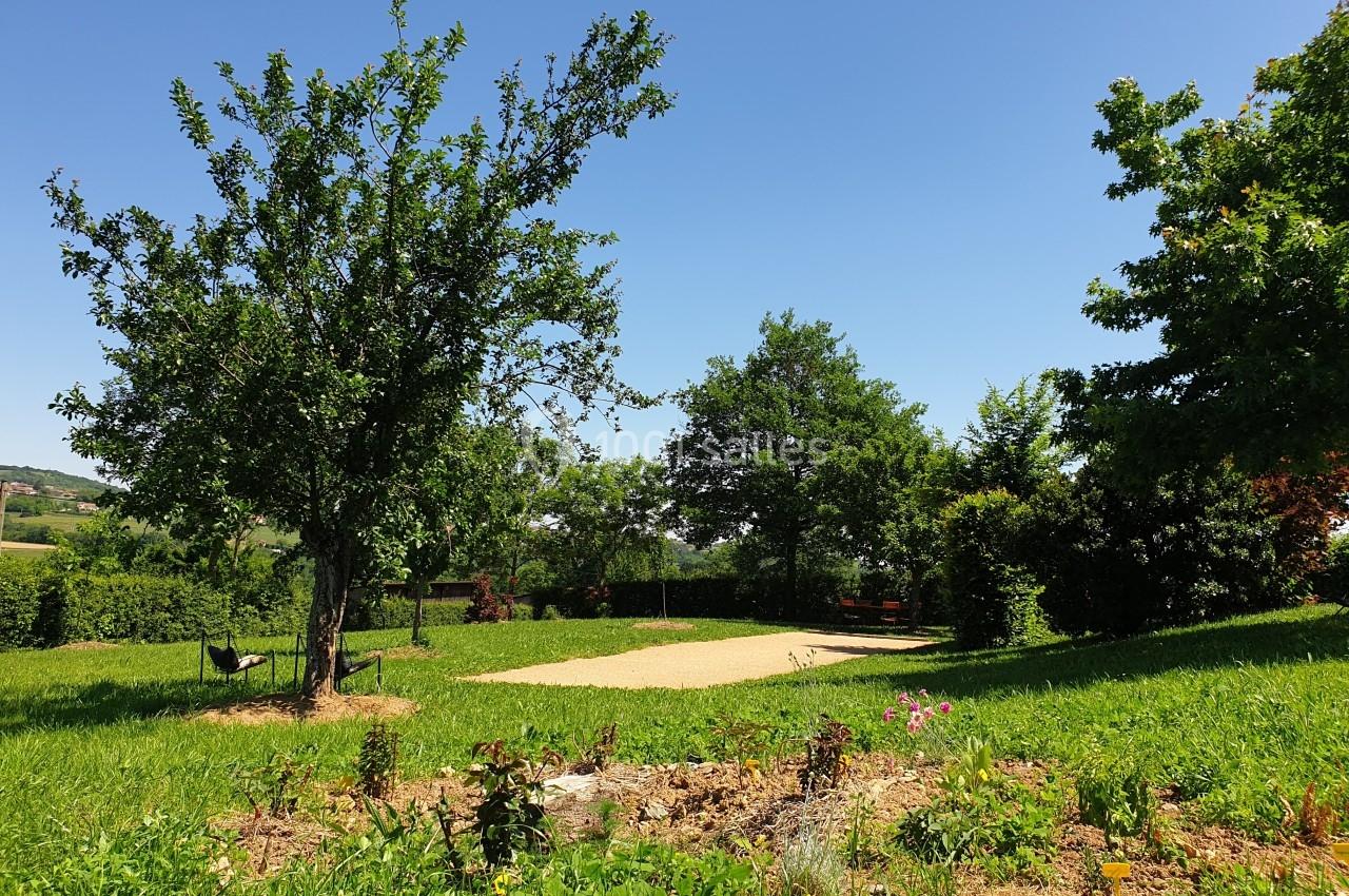 Un jardin verdoyant avec des arbres, une pelouse, des fleurs et un chemin de gravier sous un ciel bleu clair.