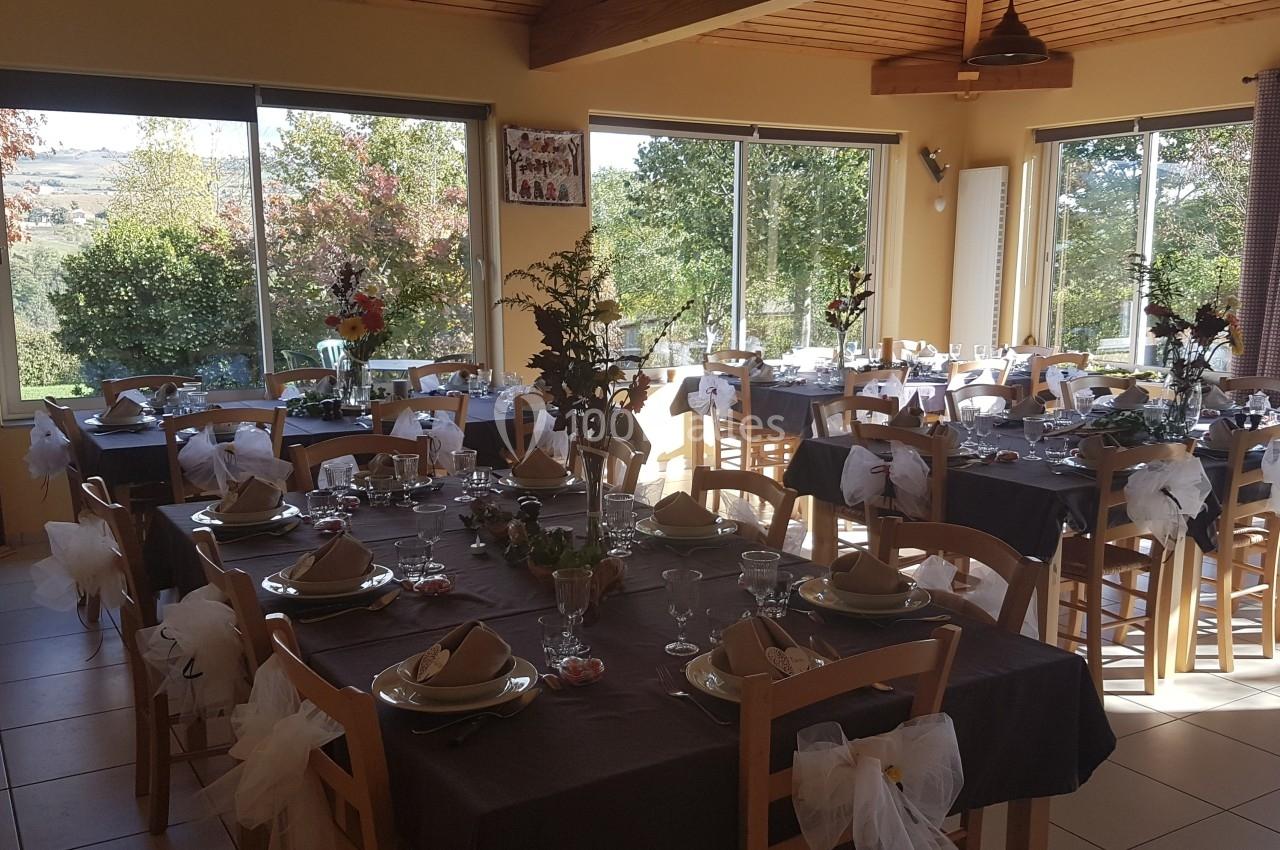 Salle lumineuse décorée pour un événement, avec des tables dressées, nappes sombres et chaises ornées de nœuds blancs.