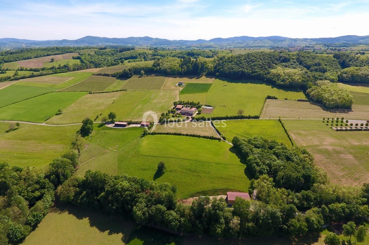 Vue aérienne d'une campagne verdoyante avec champs, arbres, bâtiments et collines à l'horizon sous un ciel dégagé.