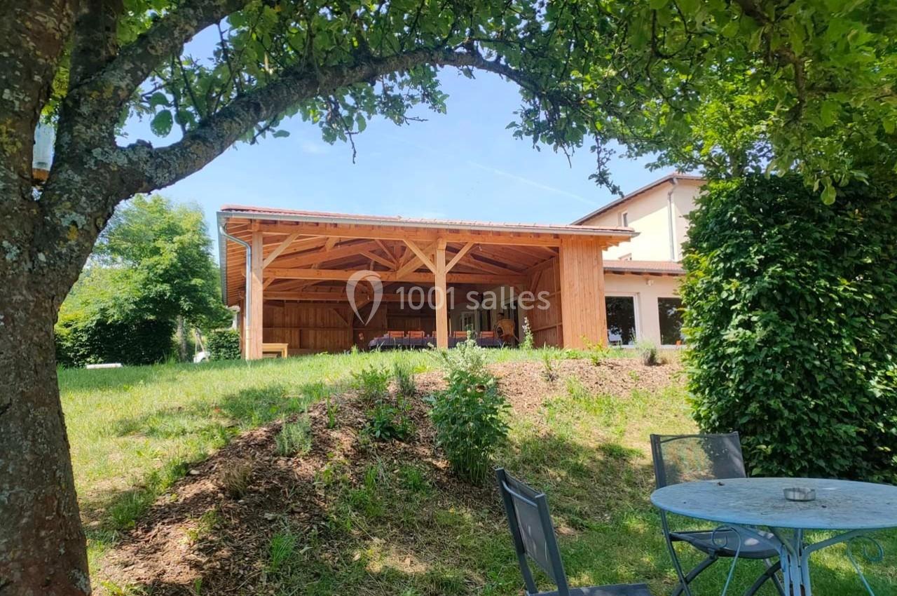 Abri en bois ouvert dans un jardin verdoyant, avec table et chaises en métal à l'ombre d'un arbre au premier plan.