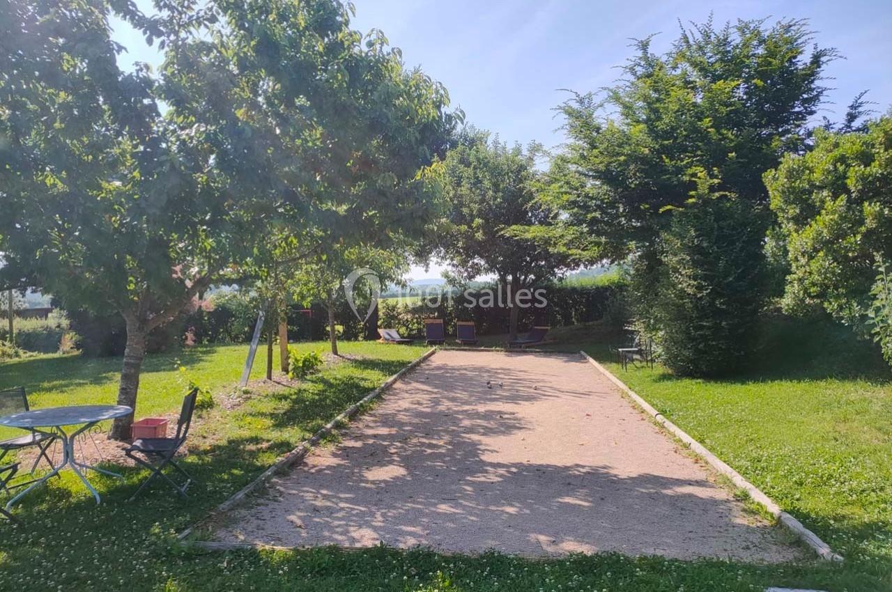 Terrain de pétanque ensoleillé entouré d'arbres et de verdure, avec des tables et chaises à proximité.