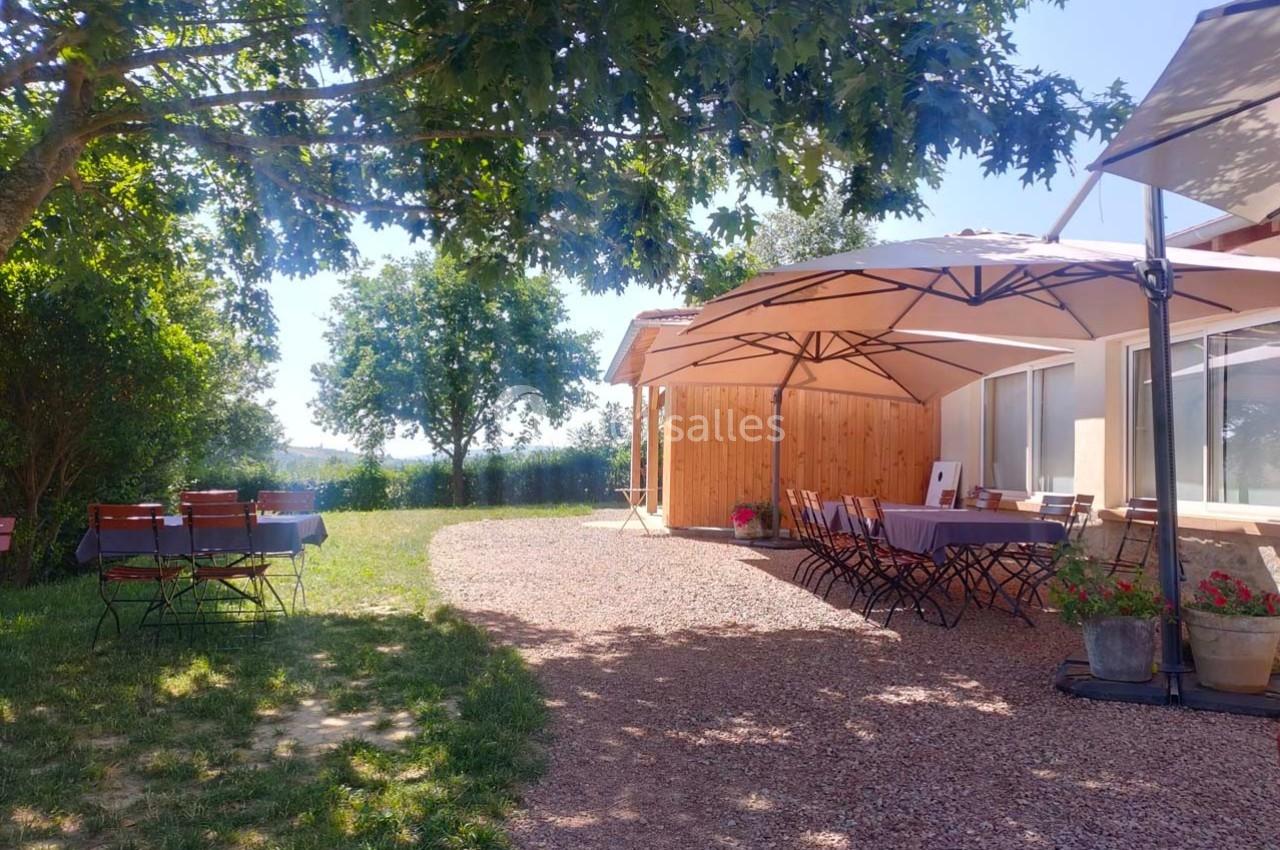 Terrasse ombragée avec tables et chaises, entourée de verdure, près d'un bâtiment en bois sous un ciel dégagé.