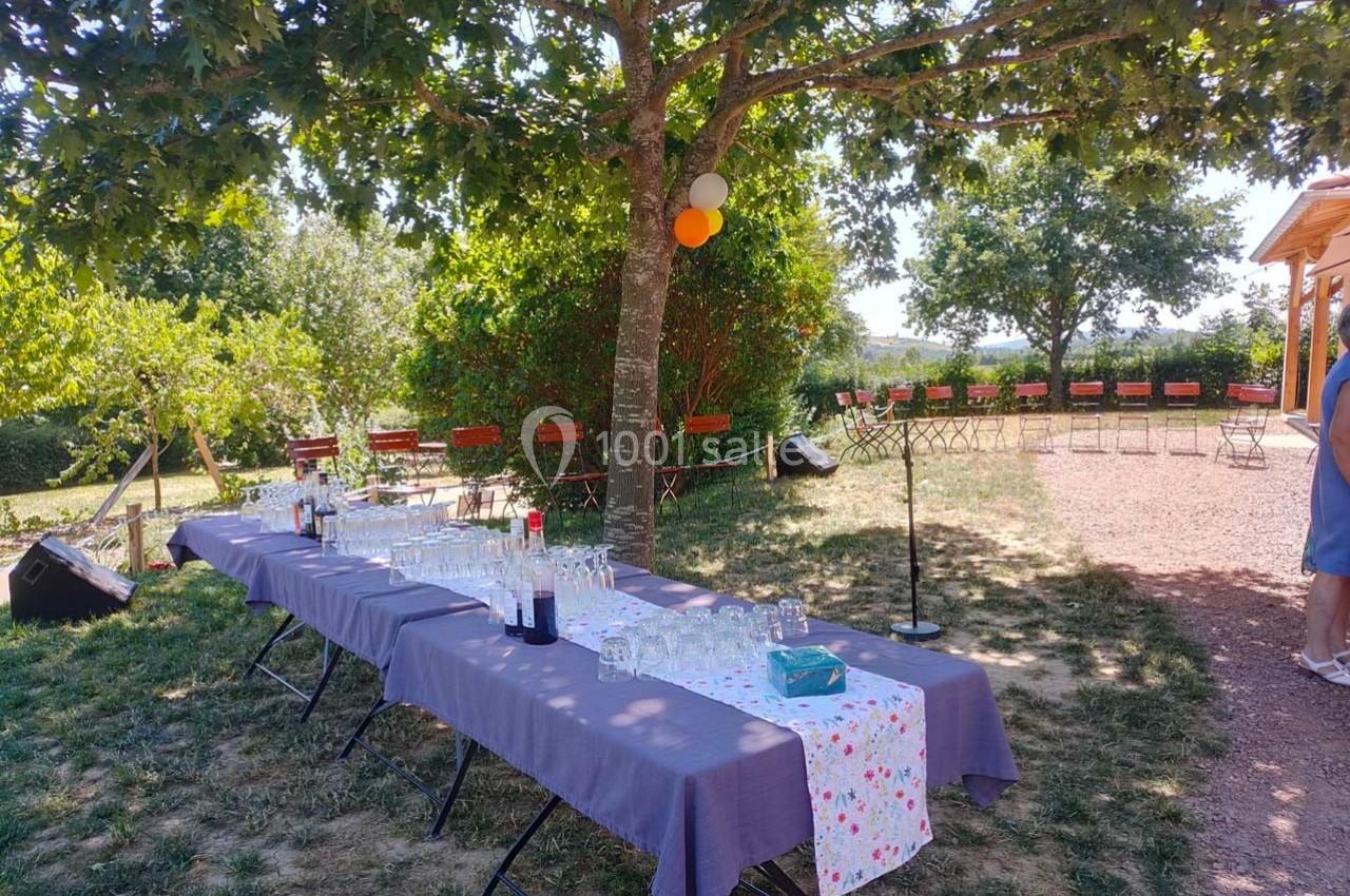 Tables dressées avec boissons et verres sous un arbre, dans un jardin avec chaises disposées en arrière-plan.