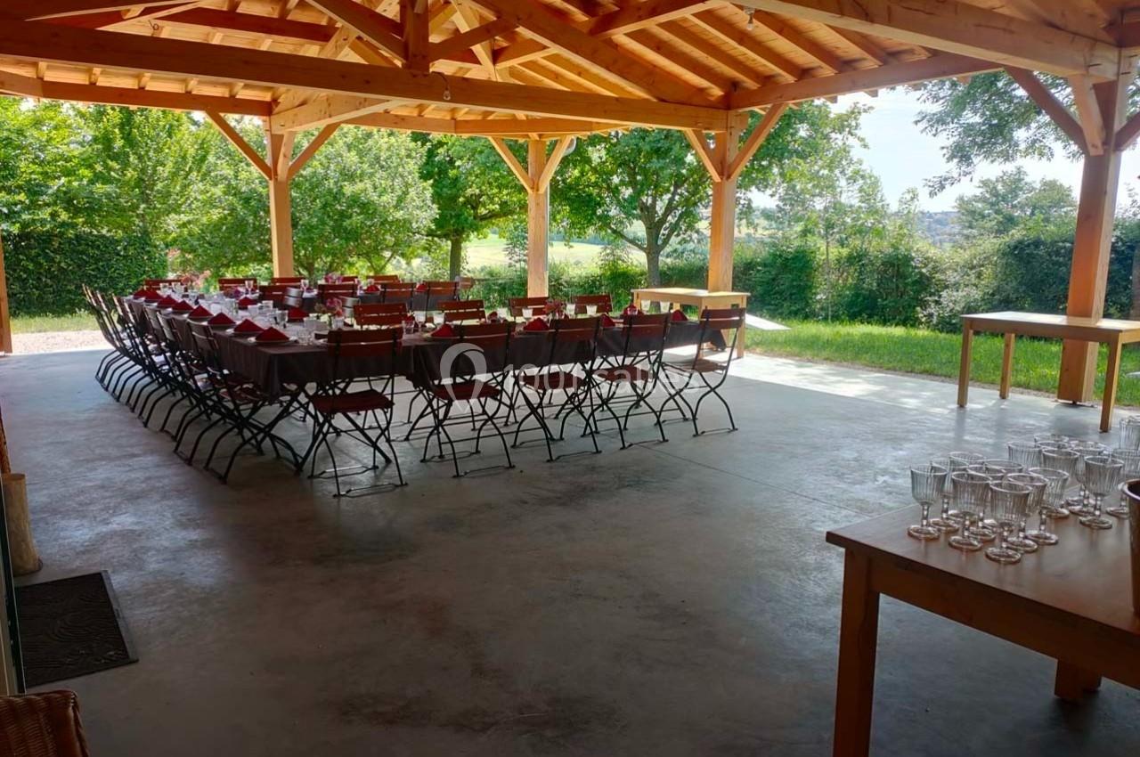 Salle extérieure couverte avec tables et chaises alignées pour un repas, entourée de verdure et éclairée par la lumière…
