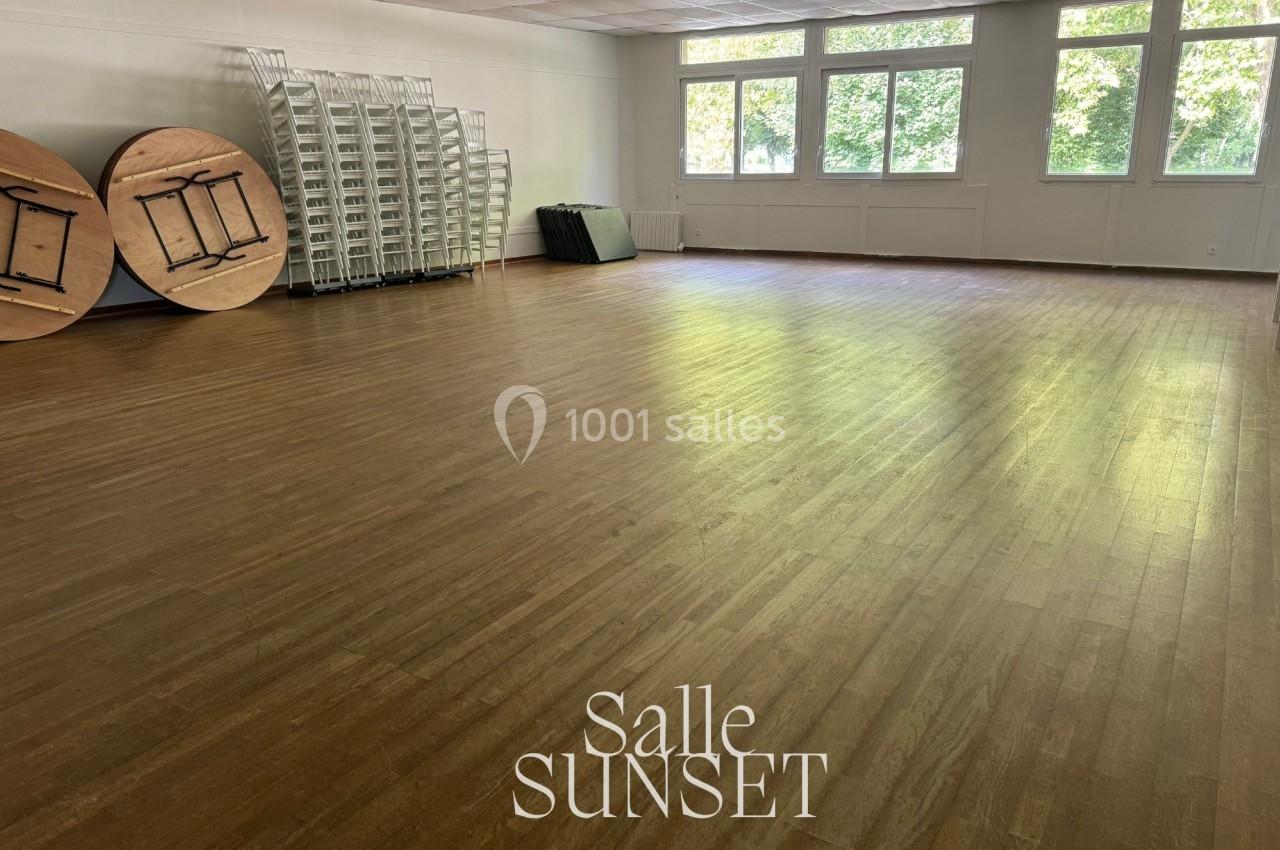 Salle vide avec parquet en bois clair, tables rondes empilées, chaises pliantes et grandes fenêtres laissant entrer la…