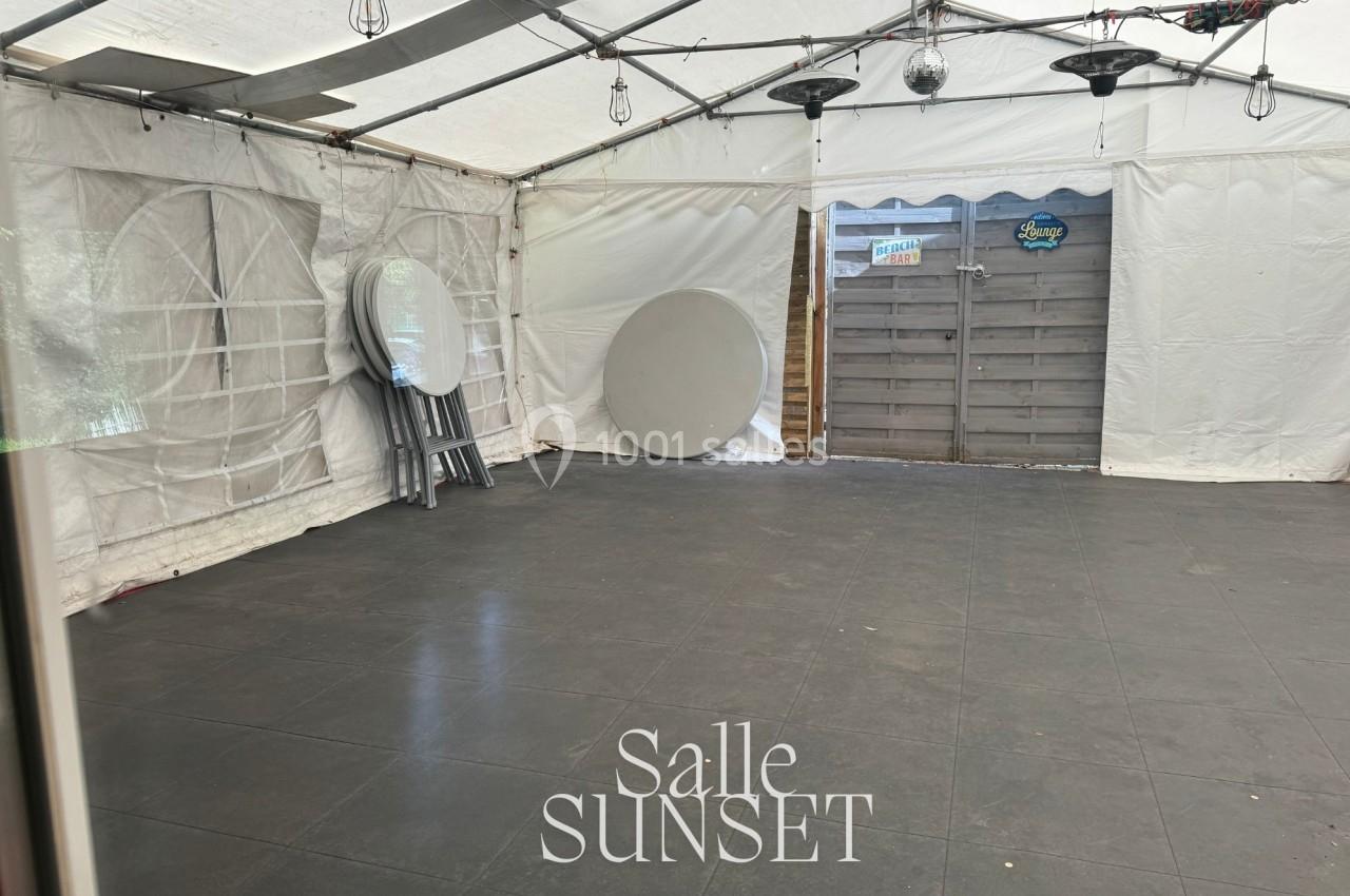 Intérieur d'une tente blanche avec sol en dalles noires, tables rondes pliées et chaises empilées sur le côté.