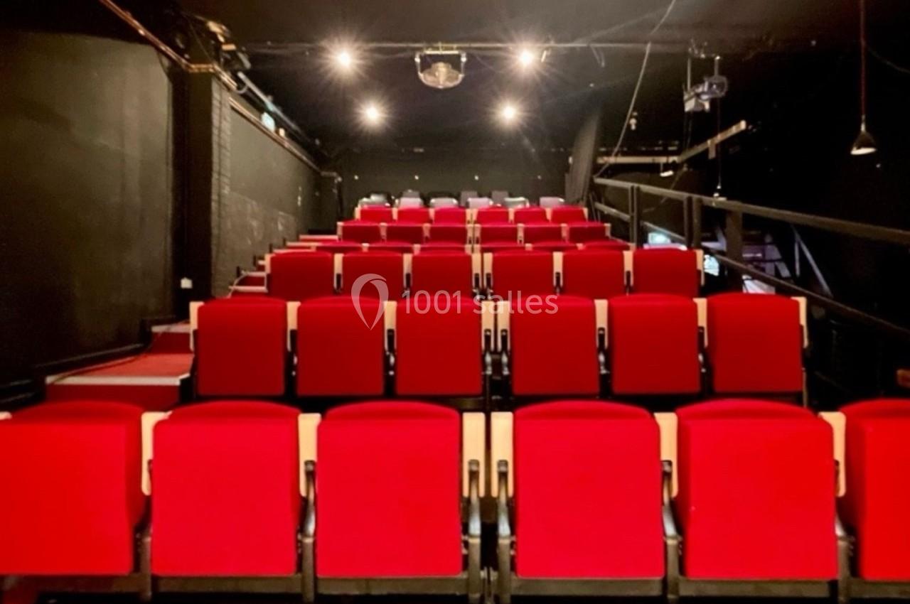 Salle de théâtre avec plusieurs rangées de sièges rouges alignés, éclairée par des spots au plafond.