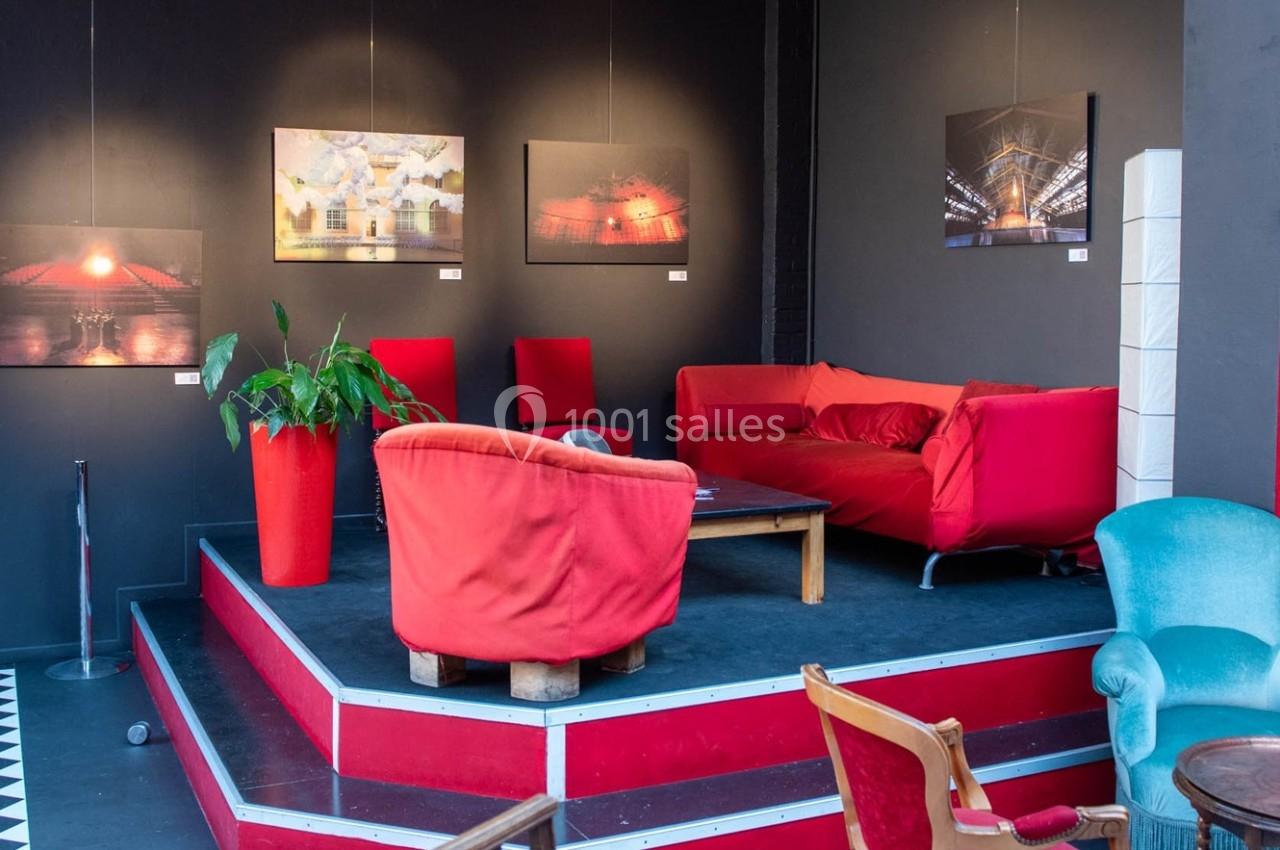 Espace salon avec canapés et fauteuils rouges, table basse en bois, plantes et photos encadrées sur un mur sombre.