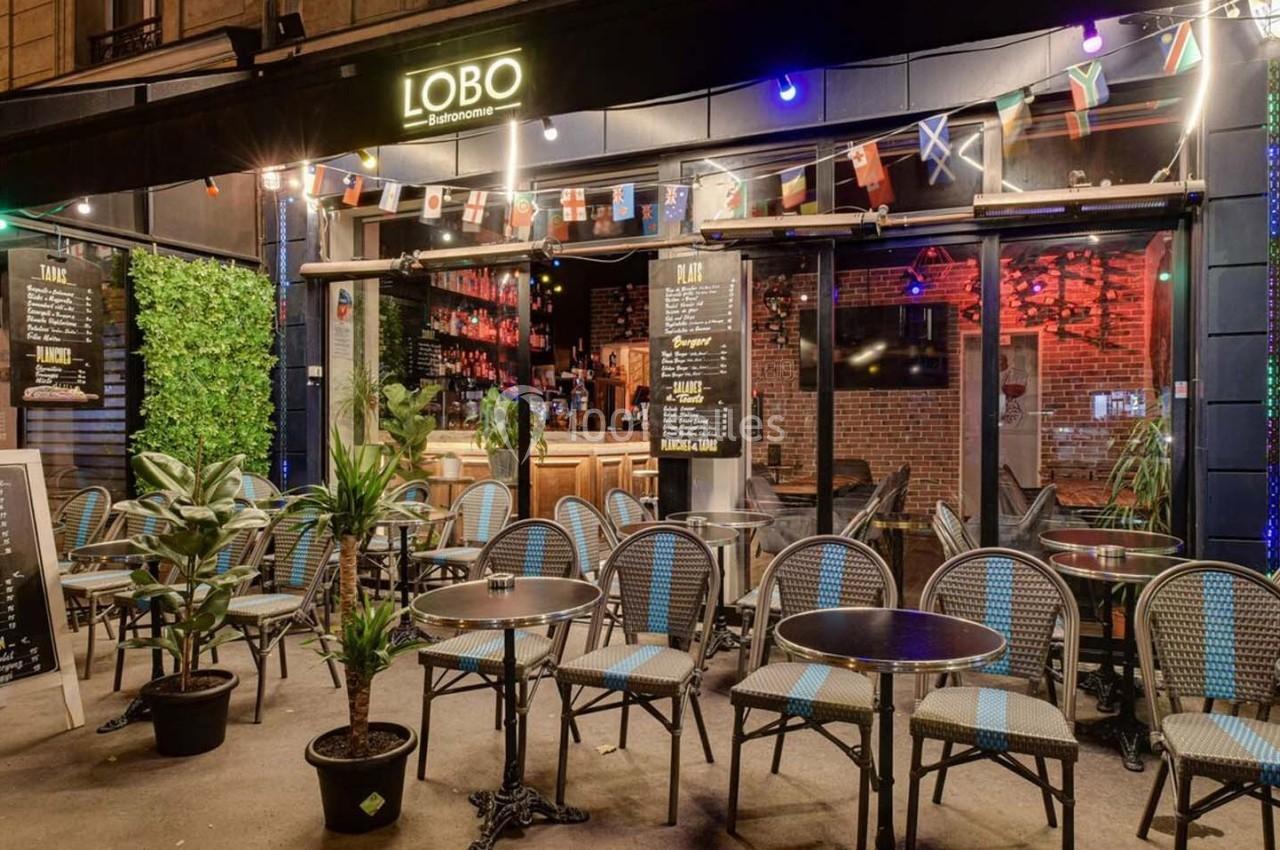 Terrasse d'un café-restaurant avec chaises en rotin, tables rondes, plantes et enseigne lumineuse ’Lobo’.
