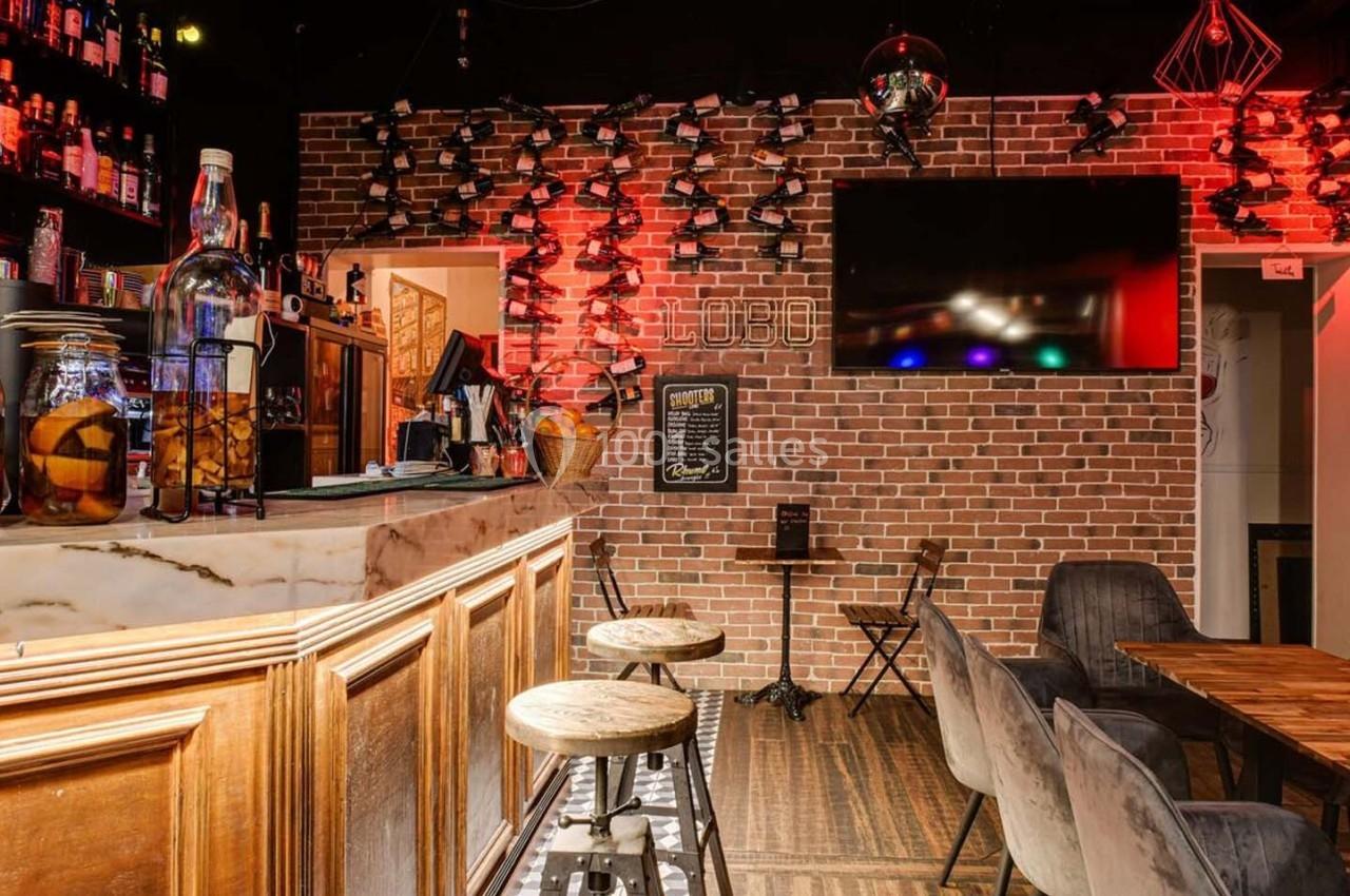 Intérieur d'un bar avec comptoir en bois, étagères de bouteilles, tables en bois et éclairage rouge tamisé.