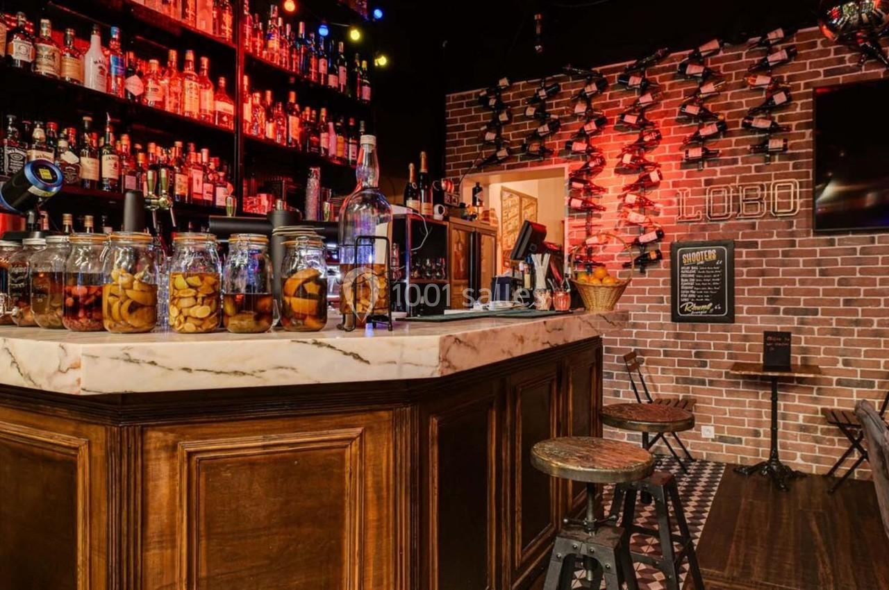 Bar avec comptoir en marbre, étagères remplies de bouteilles, bocaux de fruits et éclairage chaleureux.