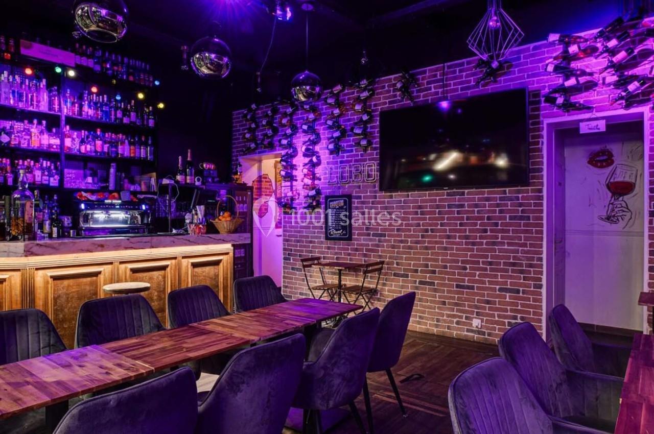Intérieur d'un bar avec comptoir en bois, étagères de bouteilles, tables en bois, chaises noires et éclairage coloré.