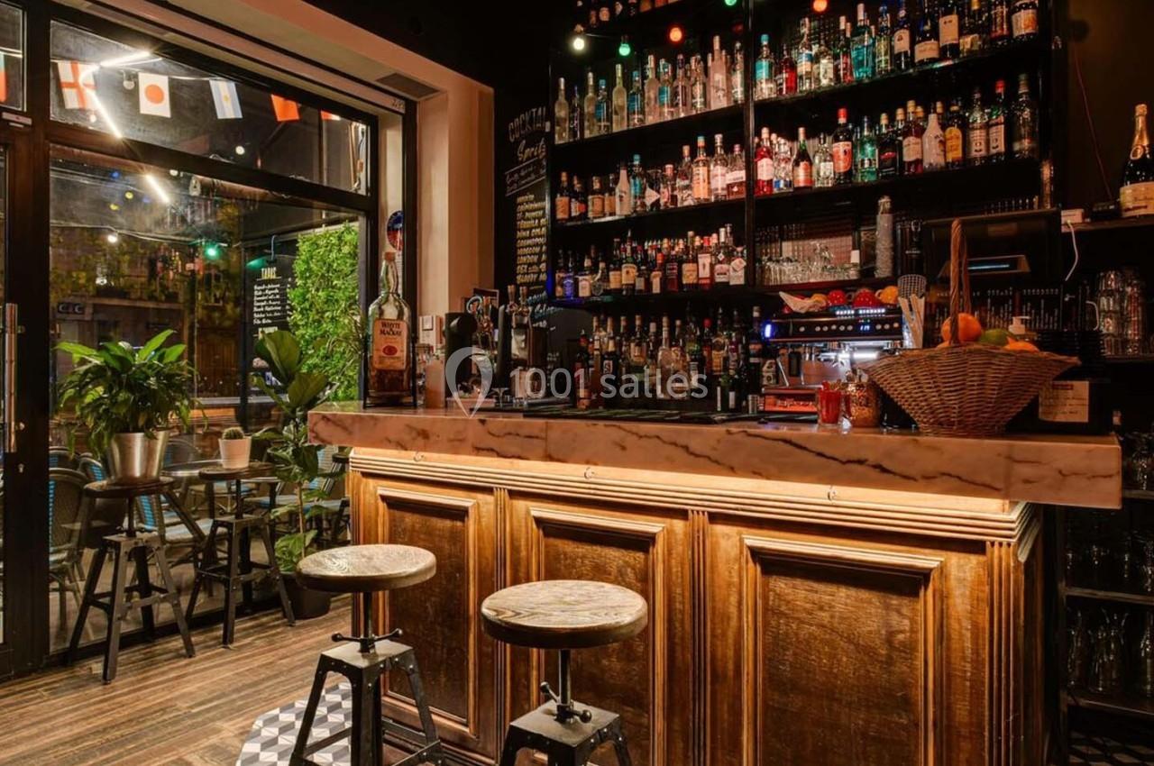 Bar chaleureux avec comptoir en bois, étagères remplies de bouteilles et tables dans un espace éclairé et décoré.