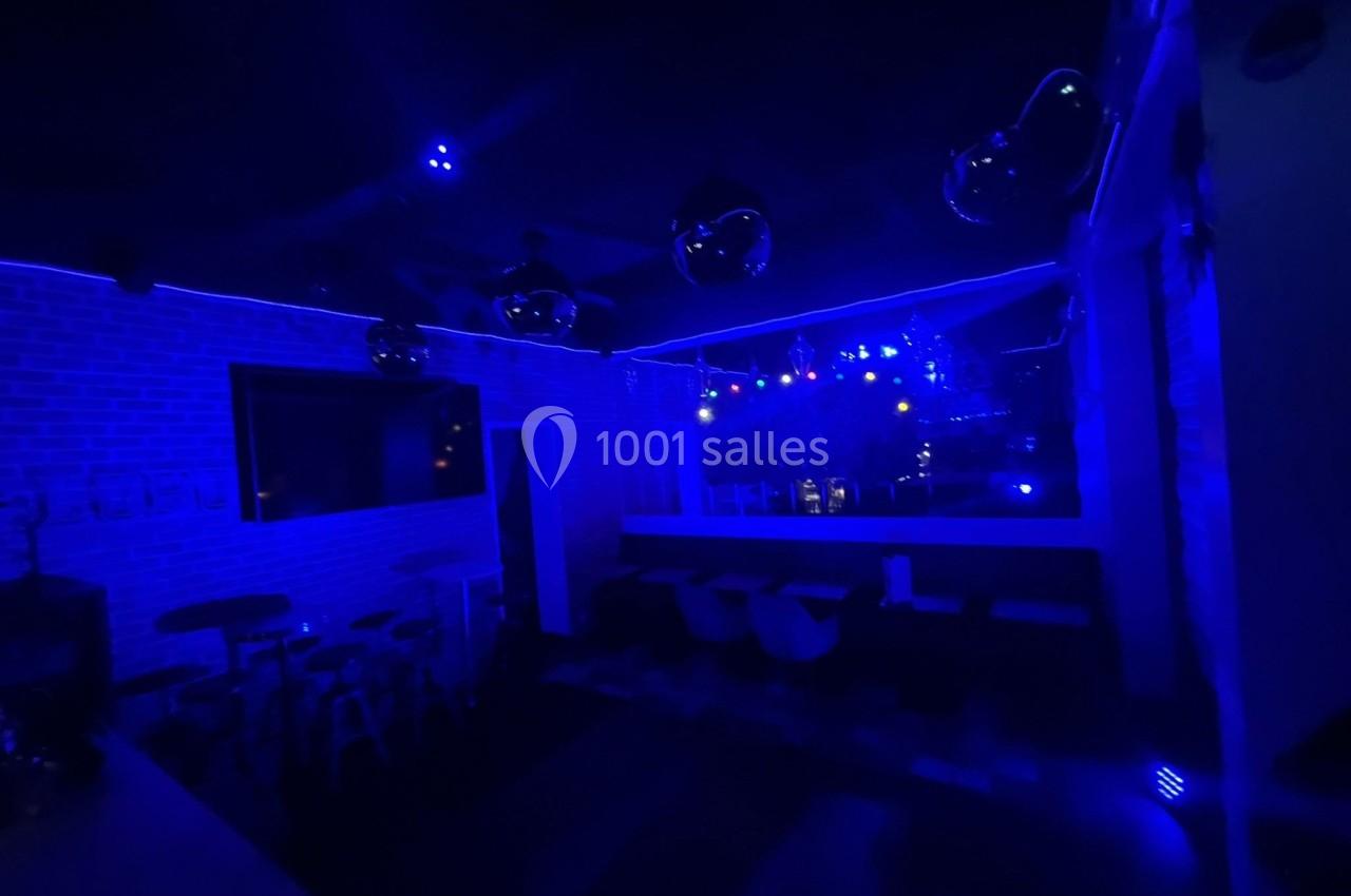 Salle sombre éclairée par des lumières bleues, avec des tables, des chaises et des murs en briques visibles.