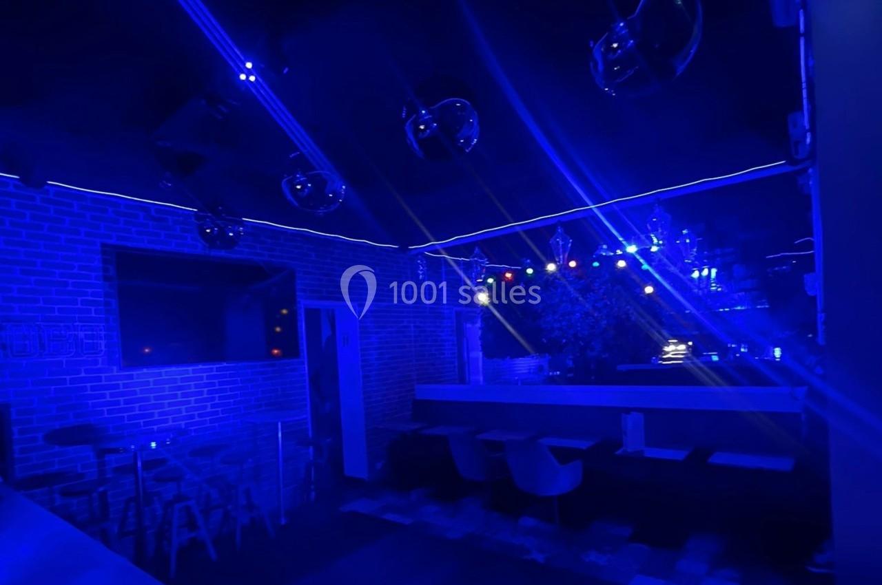 Salle sombre éclairée par des lumières bleues et colorées, avec des tables, des chaises et un mur en briques.