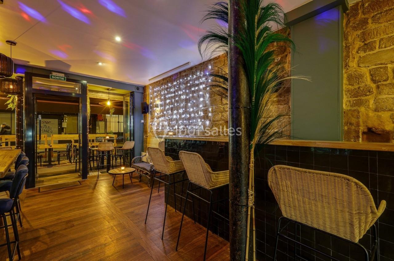Intérieur d'un bar avec des tables en bois, des chaises en rotin, un mur en pierre et des lumières décoratives.