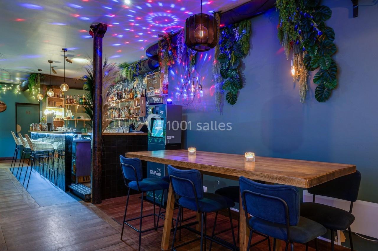 Salle de bar moderne avec un long comptoir, table en bois, chaises bleues et éclairage coloré décoratif.