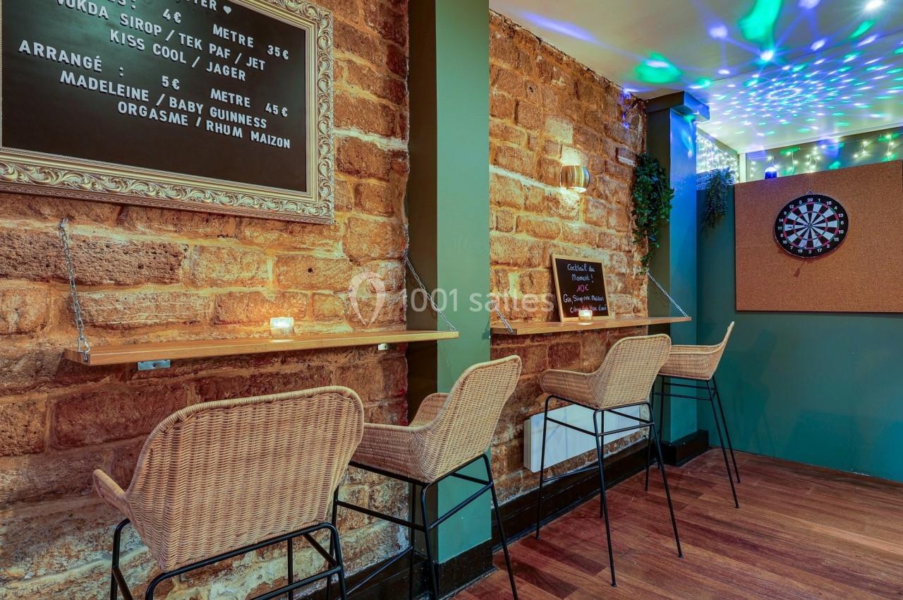 Mur en pierre avec un bar étroit, tabourets en rotin, tableau noir avec menu et cible de fléchettes au fond.