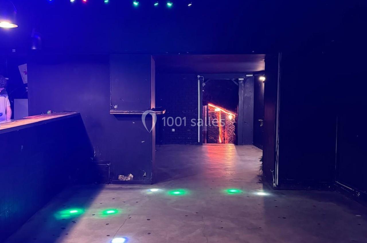 Salle sombre avec un comptoir à gauche, un sol en béton éclairé par des lumières colorées et une sortie illuminée au fond.