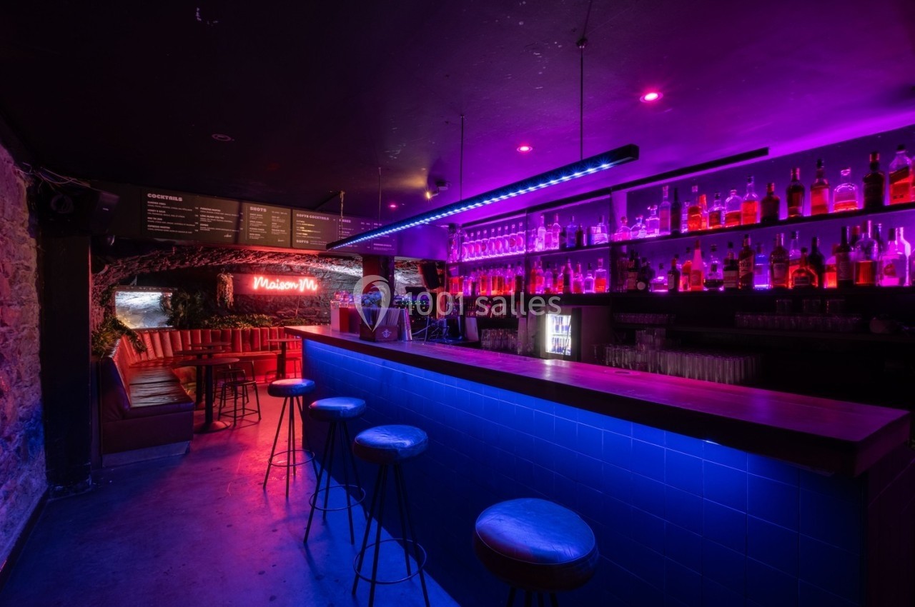 Bar moderne avec éclairage néon violet et bleu, comptoir en bois, étagères remplies de bouteilles et banquettes en arrière…