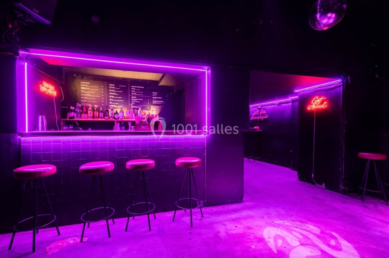 Bar avec tabourets alignés, éclairage néon violet et menu visible en arrière-plan dans une ambiance sombre.