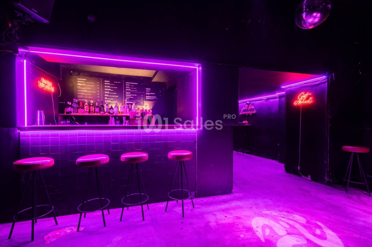 Bar avec tabourets alignés, éclairage néon violet et menu visible en arrière-plan dans une ambiance sombre.