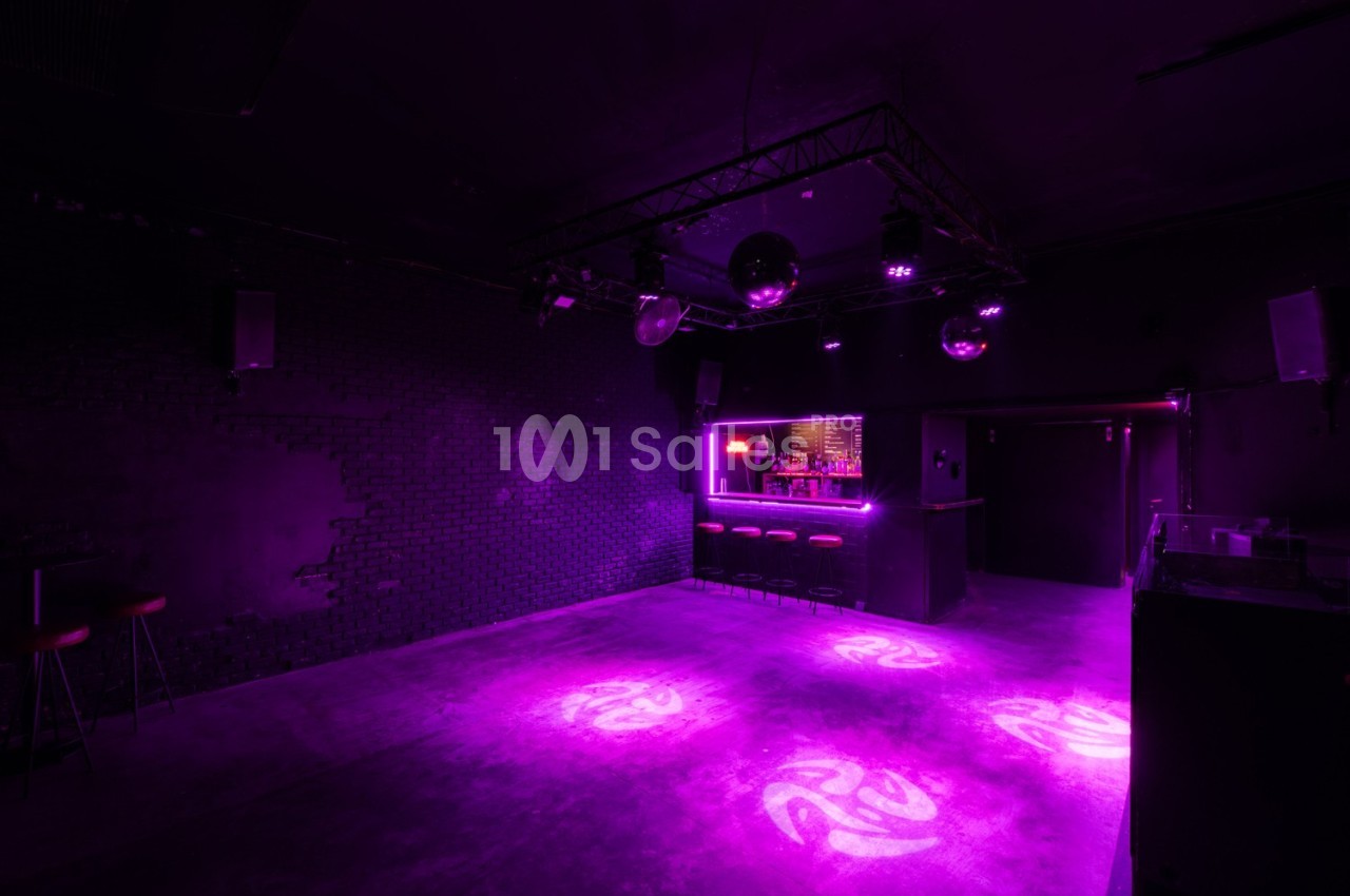 Salle de danse vide avec éclairage violet, bar illuminé en arrière-plan et murs sombres.