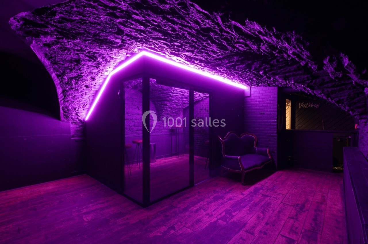 Salle voûtée avec éclairage néon violet, parquet en bois et mobilier sombre, comprenant un canapé et une cabine vitrée.