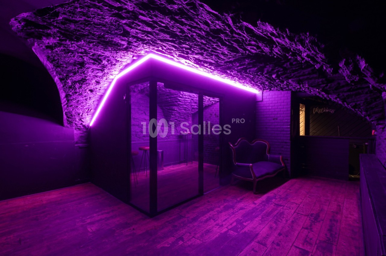 Salle voûtée avec éclairage néon violet, parquet en bois et mobilier sombre, comprenant un canapé et une cabine vitrée.