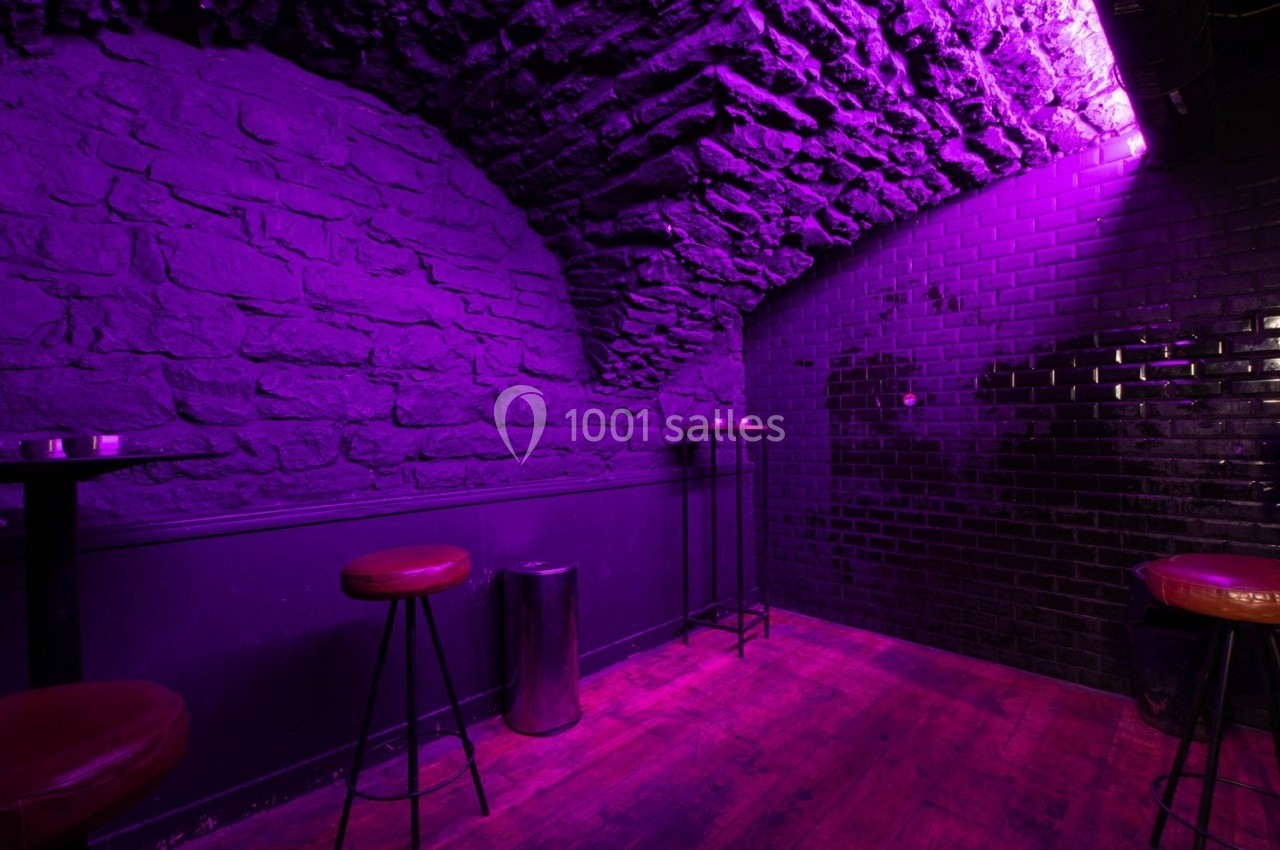 Salle voûtée en pierre éclairée par une lumière violette, avec tabourets hauts, tables et sol en bois.