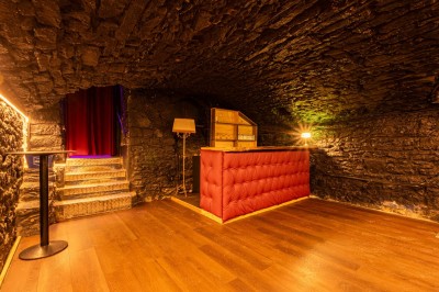 Miniature Speakeasy privatisable Salle voûtée en pierre avec un comptoir rouge, éclairage chaleureux et escalier menant à un rideau rouge.